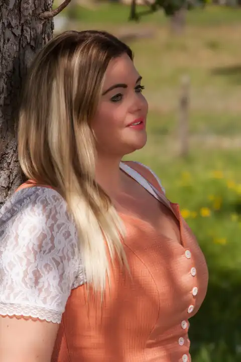 The picture shows a plus-size model with long blonde hair leaning against a picturesque tree. The model wears a striking orange dirndl and exudes confidence as she gazes dreamily into the camera. The natural beauty of the surroundings and the striking presence of the model merge to create a romantic scene.