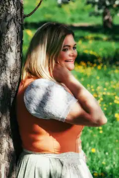 The picture shows a plus-size model with long, blonde hair smiling and leaning against an idyllic tree. One hand gently plays with the model's hair as she looks into the camera with a beaming smile. The model wears a striking orange dirndl that emphasizes her voluptuous curves and radiates self-confidence and naturalness.