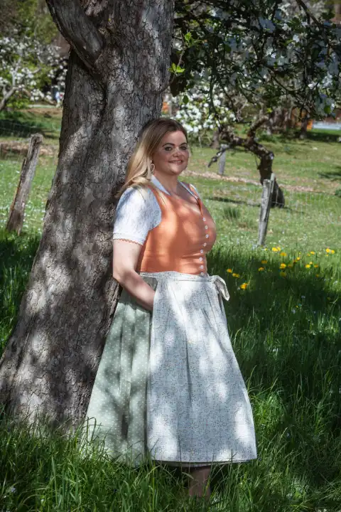 In this appealing image, a blonde plus-size model with a beaming smile on her lips stands by a picturesque tree. The model's hands rest relaxed in her pockets as she looks into the camera with natural grace and composure. The eye-catching orange dirndl gently hugs the model's voluptuous curves and emphasizes her feminine beauty. The long blonde hair frames the face and lends the portrait a timeless elegance.