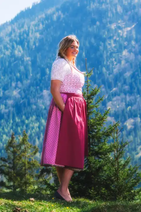 The picture shows a radiant plus-size model standing in a picturesque mountain meadow in the Alps. With her hands casually hidden in her pockets and long blonde tresses, the model presents herself in an enchanting pink dirndl. The majestic mountain backdrop and the model's charming smile lend the scene a special grace and naturalness.