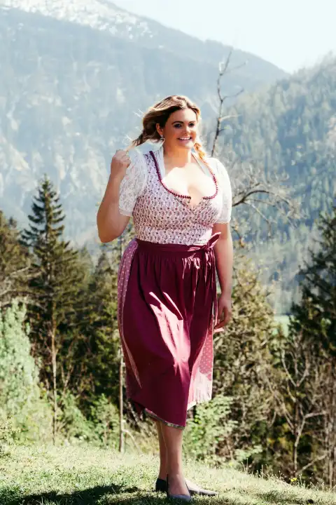 Eine junge Frau mit strahlendem Lächeln und blonden Zöpfen präsentiert sich im traditionellen bayerischen Dirndl. Das Kleid betont ihre üppigen Kurven und unterstreicht ihre feminine Ausstrahlung. Ihre fröhliche Art und ihr selbstbewusstes Auftreten machen sie zu einem Blickfang.