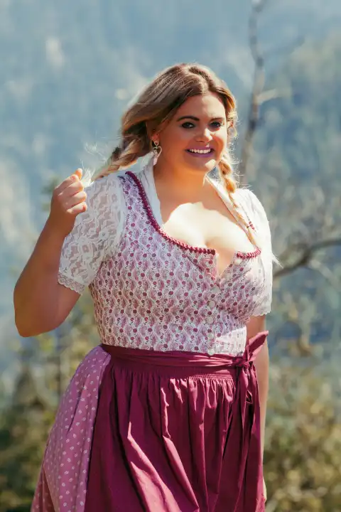 Eine junge Frau mit strahlendem Lächeln und blonden Zöpfen präsentiert sich im traditionellen bayerischen Dirndl. Das Kleid betont ihre üppigen Kurven und unterstreicht ihre feminine Ausstrahlung. Ihre fröhliche Art und ihr selbstbewusstes Auftreten machen sie zu einem Blickfang.
