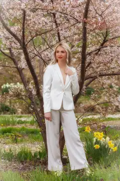 A young, fashionable blonde woman stands under a blossoming tree. She is wearing an elegant white trouser suit and holding a pair of sunglasses in her hand. The scene radiates freshness and spring.
