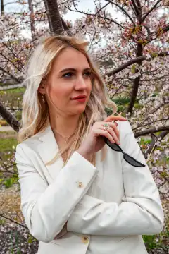 A portrait of a young, blonde woman holding a pair of sunglasses. In the background is a flowering shrub, creating a spring-like and vibrant mood. The woman radiates self-confidence and style, and the interplay of natural beauty and fashionable accessory gives the image a special touch.