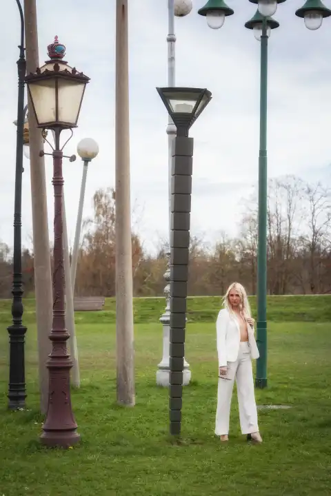 An attractive, blonde woman in a white, fashionable trouser suit stands in the middle of a meadow, surrounded by various street lamps. Her stylish outfit and confident pose stand out against the unusual background, creating an image full of contrasts and modern aesthetics.