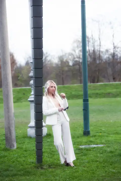 An attractive, blonde woman leans against a lamppost in a white trouser suit with an open jacket that shows her chest. She is in the middle of a meadow, which creates an interesting contrast between urban and natural surroundings. Her confident expression and fashionable outfit emphasize her elegance and style.