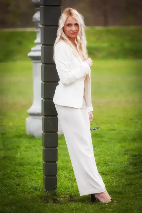 A young, blonde woman in a white trouser suit leans against a lamppost in the middle of a meadow and looks directly into the camera. Her confident posture and fashionable outfit create a strong visual appeal and emphasize her modern elegance and style.