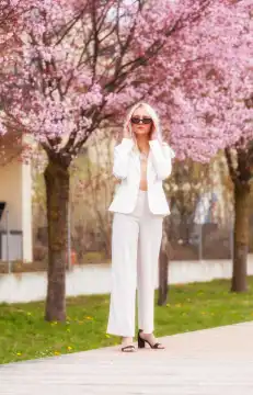 Eine stilvolle Frau im weißen Hosenanzug steht vor einem blühenden Baum und setzt ihre Sonnenbrille auf. Die Szene vereint natürliche Schönheit mit modischem Flair und strahlt Eleganz und Selbstbewusstsein aus.