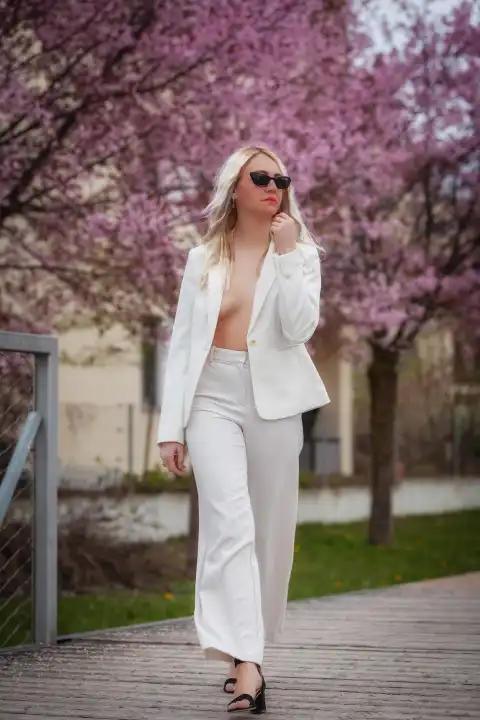 A model wears a white trouser suit with an open jacket that shows off her chest and walks through a park full of blossoming trees. She wears sunglasses and exudes elegance and self-confidence as she enjoys the springtime surroundings.