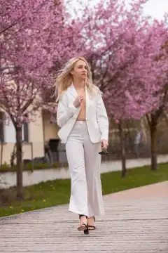 The young woman wears a white trouser suit and holds a pair of sunglasses in her hand as she walks through a park full of blossoming trees.