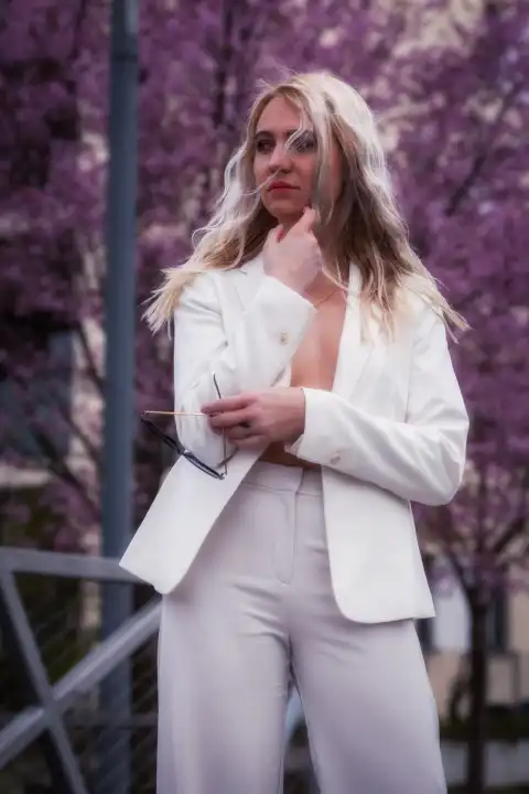 The blonde woman is standing in a 3/4 shot, wearing a white trouser suit and has her jacket open.