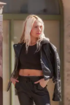 The young woman stands in a 3/4 shot and wears a trendy leather outfit with a belly shirt that emphasizes her fashion affinity and self-confidence.