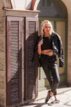 Das blonde junge Fotomodel lehnt sich in einem verführerischen Lederoutfit in einer sexy Pose an die Mauer eines Cafés und strahlt Selbstbewusstsein aus.