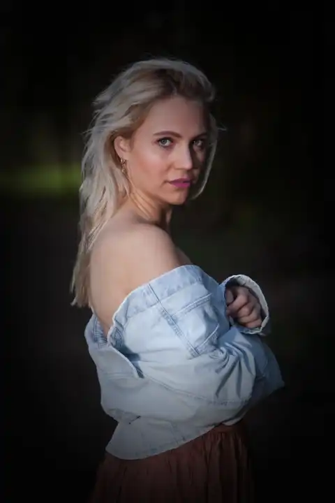 Eine Oberkörperaufnahme einer jungen blonden Frau, die lässig eine Jeansjacke trägt und ihre nackten Schultern zeigt. Sie blickt direkt in die Kamera, was Intimität und Selbstbewusstsein ausstrahlt.