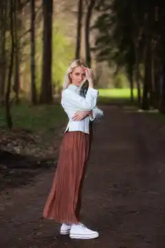 A full-body shot shows a young blonde woman standing casually in the forest. She is wearing a denim jacket, a long brown skirt and sneakers, creating a casual yet stylish look.