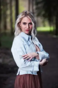 In an upper body shot, a young blonde woman stands in the forest. She is wearing a denim jacket and looks confidently straight into the camera, while the sunlight falls through the trees and creates a warm atmosphere.