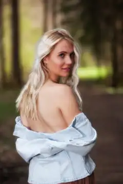 In an upper body shot, a young blonde woman stands in the forest. Her denim jacket is pulled down, exposing her bare shoulders. She looks intensely and confidently directly into the camera, while the light falls through the trees and creates a magical atmosphere.