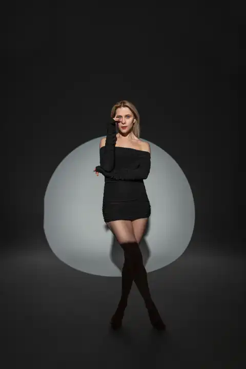 Mode schöne stilvolle Mädchen Modell in einem schwarzen sexy Kleid mit schlanken Beinen steht und posiert auf einem dunklen Hintergrund in einem Studio mit kreisförmigen Licht
