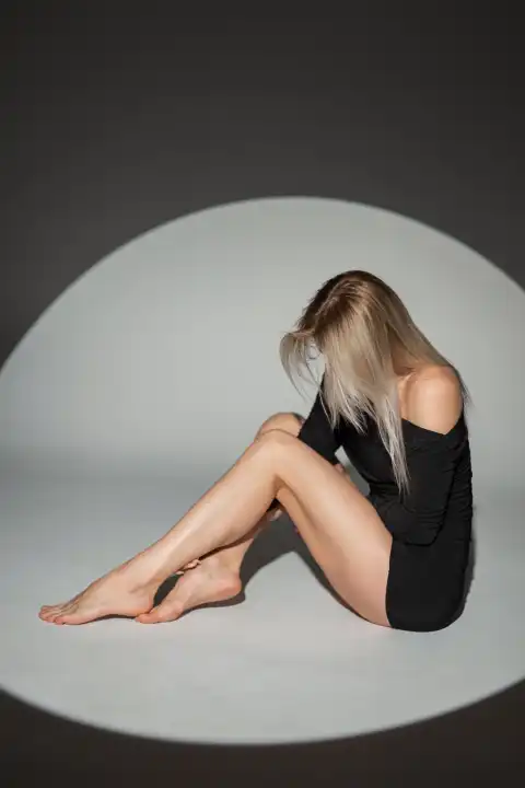 Mode schöne stilvolle blonde Mädchen mit einem schlanken Körper und Beine in einem schwarzen sexy Kleid sitzt in einem Studio mit kreisförmigen Licht
