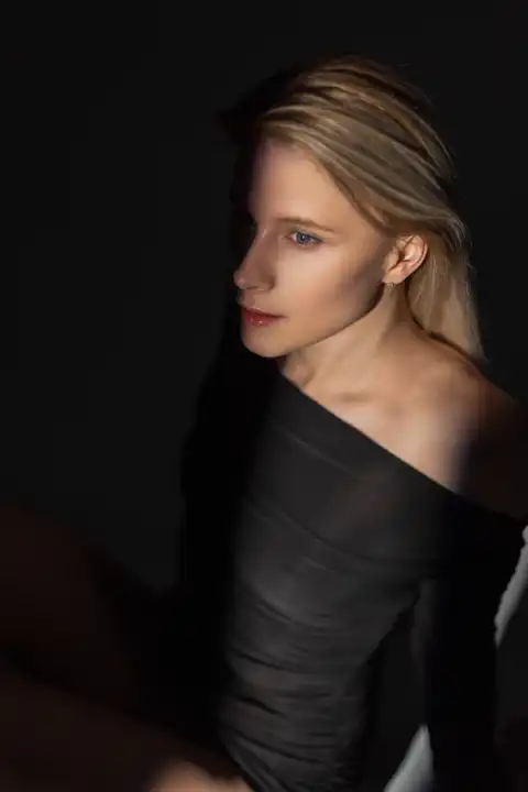 Studio portrait of a beautiful fashion blond woman in a black dress sitting in the dark with a ray of light
