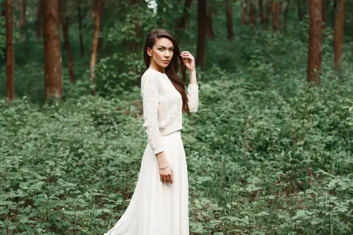 Outdoors portrait of beautiful young caucasian brunette woman in white dress over green foliage on background