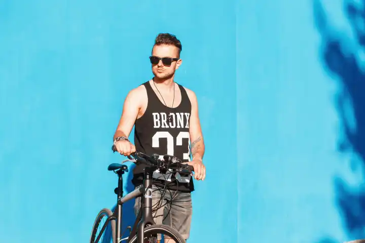 Hipster-Mann mit Sonnenbrille auf einem Fahrrad in der Nähe einer hellblauen Wand.