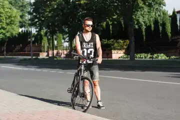Junge stilvolle Mann steht auf der Straße mit dem Fahrrad auf dem Hintergrund der Grünanlage.