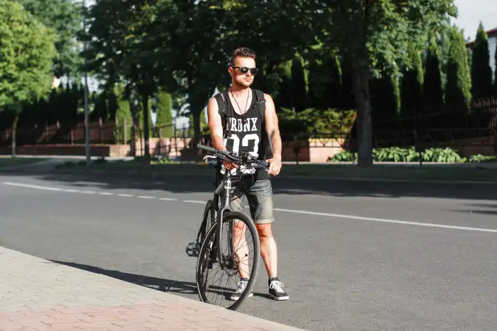 Junge stilvolle Mann steht auf der Straße mit dem Fahrrad auf dem Hintergrund der Grünanlage.
