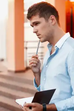 Junger Mann mit Notizbuch und Stift, der über seine Arbeit nachdenkt.