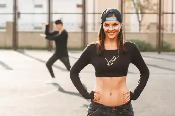 Hip-hop dancer woman in stylish black dress with chain "Loves" and a cap. Man standing in a dancing pose on the background.