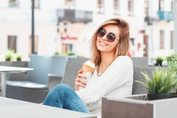 Modische schöne lächelnde Frau mit runder Sonnenbrille und Eiscreme, die sich in einem Sommercafé entspannt.