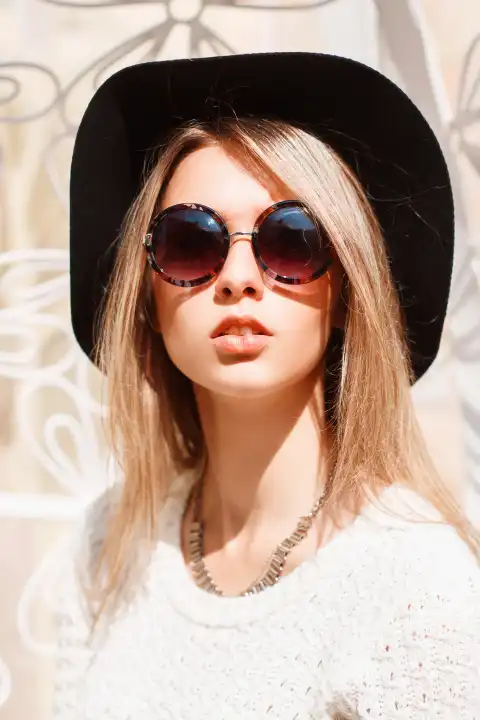 Portrait of a beautiful young girl in a black hat and round sunglasses on a sunny day.
