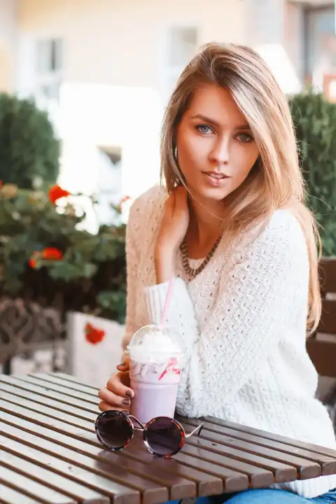 Hübsche Frau im weißen Pullover sitzt in einem Sommercafé und trinkt einen Cocktail