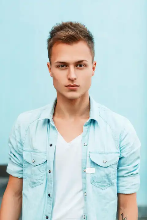 Close-up portrait of a young stylish guy in a denim shirt near a blue wall.