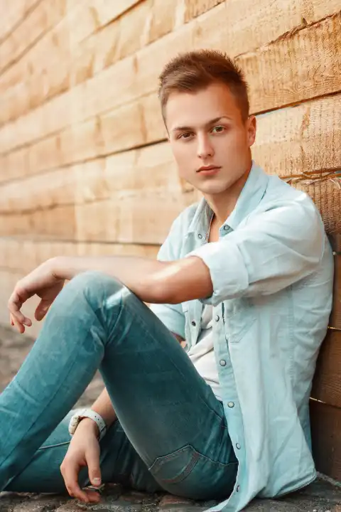 Junger gut aussehender Mann in Jeanskleidung sitzt an einer Holzwand.