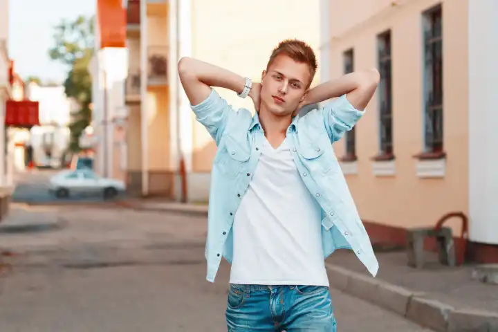 Young handsome man in a denim shirt and jeans holding hands behind head on the background of the city.
