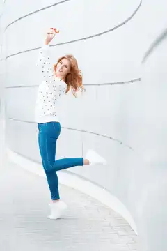 Beautiful girl with a smile in a white sweater, blue jeans and white sneakers jumping. Happy outdoor portrait on the background of modern metal wall.