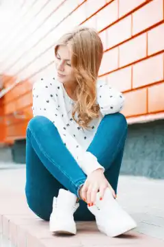 Schöne Frau in einem weißen Pullover, Jeans und weißen Turnschuhen sitzt auf einer Fliese in der Nähe der orangefarbenen Wand