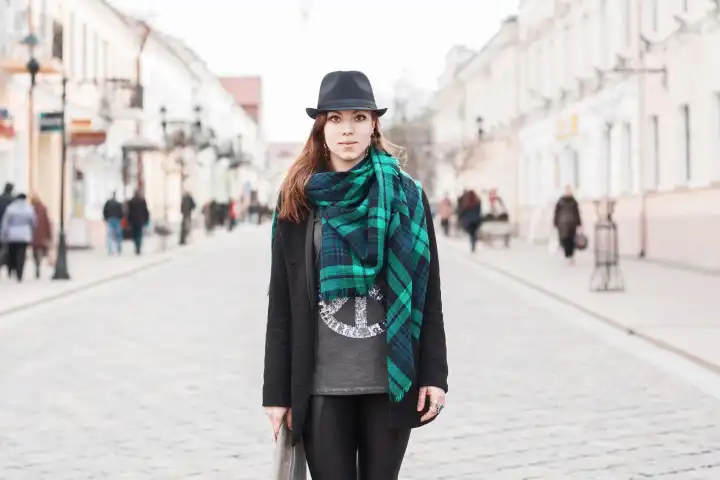 Portrait of a beautiful girl in a black cloak with a scarf standing on the square. People walking around.
