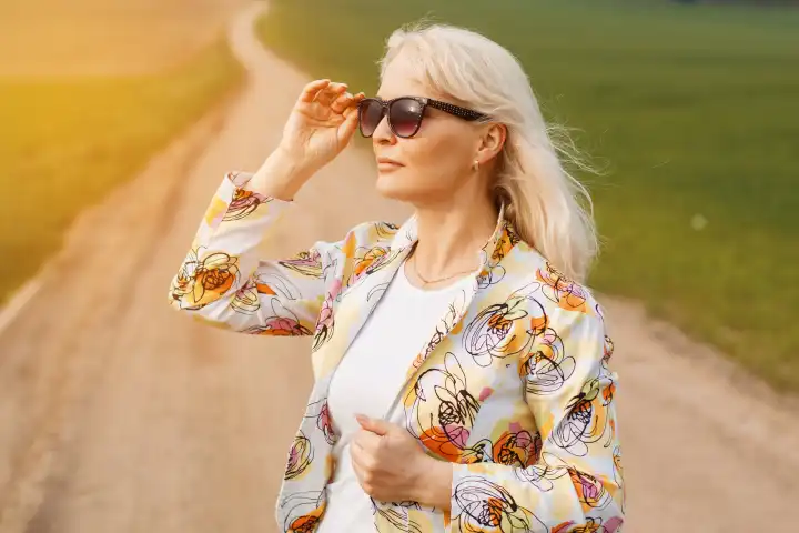 Ältere Frau in einer Jacke auf der Straße auf der grünen Wiese. Hält eine Sonnenbrille.