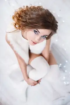 Pretty woman with curls in a fur sleeveless jacket and knitted stockings sitting on the white wooden floor. Top view.