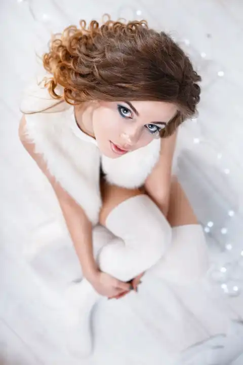 Hübsche Frau mit Locken in einer ärmellosen Pelzjacke und gestrickten Strümpfen sitzt auf dem weißen Holzboden. Ansicht von oben.