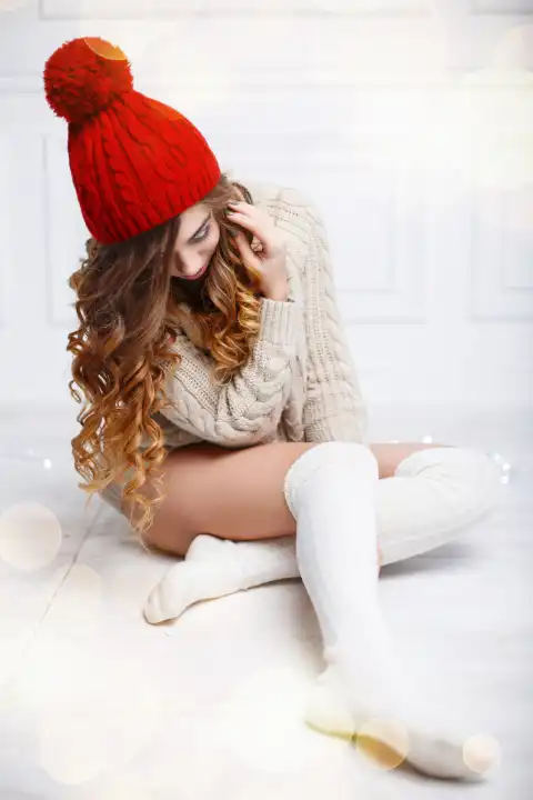 Beautiful fashionable American girl in knitted warm clothes on a background of lights. Woman sitting on white wooden floor
