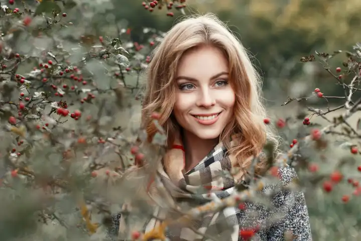 Schönes junges Mädchen mit einem Lächeln in einem warmen Schal in der Nähe eines Baumes mit Beeren