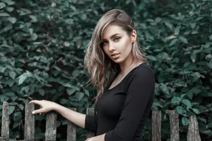 Schönes Mädchen in einem schwarzen Hemd steht in der Nähe des Holzzauns auf dem Hintergrund des grünen Laubes.