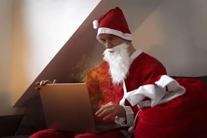 Weihnachtsmann auf dem Sofa mit einem Laptop Computer und bestellt Geschenke für Weihnachten online im Internet zur rechtzeitigen Lieferung an Heilig Abend. E-Commerce Website Weihnachten Zeit Einkauf Konzept
