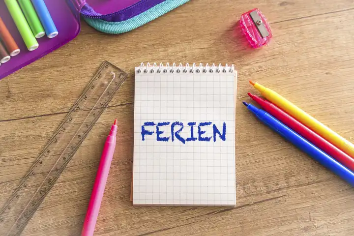 Notizblock auf einem Schultisch mit bunten Stiften mit der Aufschrift: Ferien. Schulferien Konzept FOTOMONTAGE