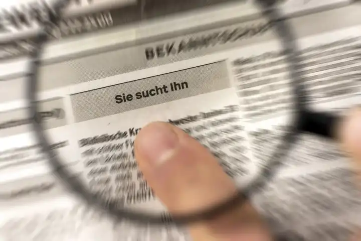 Augsburg, Bavaria, Germany - 2 April 2023: Man holding magnifying glass over daily newspaper with ads about personals section and headline: Sie sucht Ihn