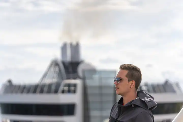Ein Mann steht auf einem Kreuzfahrtschiff. Er trägt eine Jacke und eine Sonnenbrille