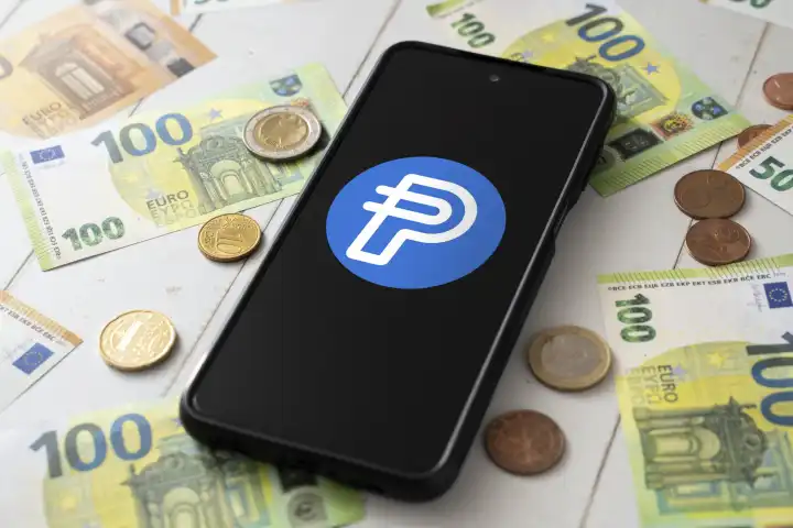 8 August 2023: Smartphone with Paypal USD logo on screen next to Euro bills and coins. Cryptocurrency PYUSD from Paypal PHOTOMONTAGE